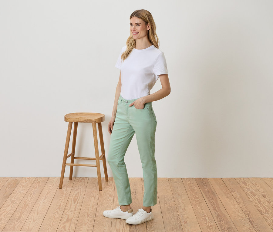 Nő áll egy zsámoly mellett, fehér pólót, zöld nadrágot és fehér tornacipőt visel.