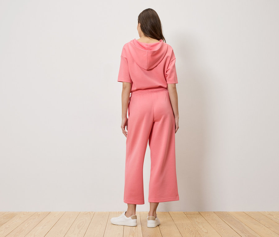 Egy nő háttal áll a kamerának, rózsaszín kapucnis pulóvert és culotte nadrágot visel.