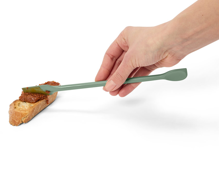 Egy kéz zöld spatulával piros krémet ken egy szelet kenyérre.