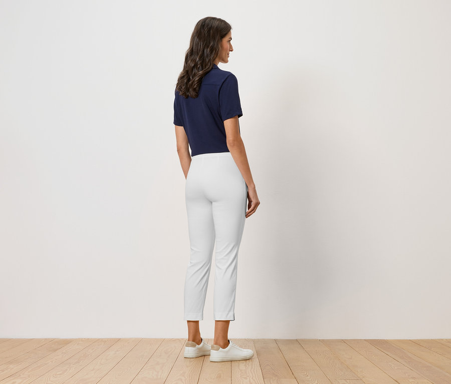 Egy nő áll háttal a kamerának, sötétkék galléros pólót, fehér nadrágot és fehér cipőt visel.