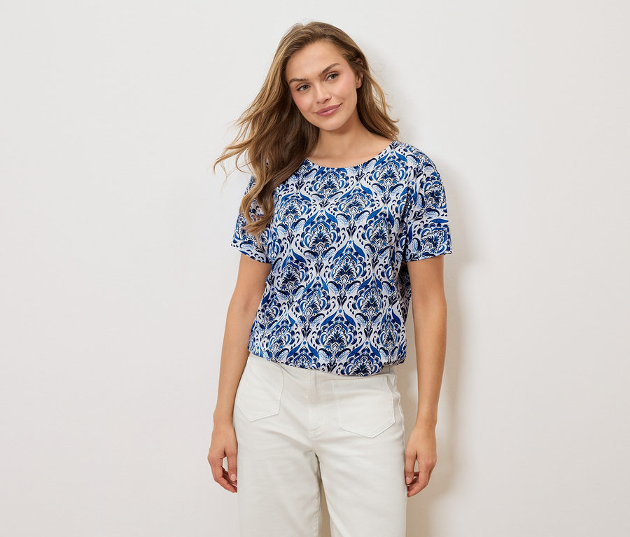 Nő visel kék-fehér mintás pólót és fehér nadrágot fehér háttér előtt.