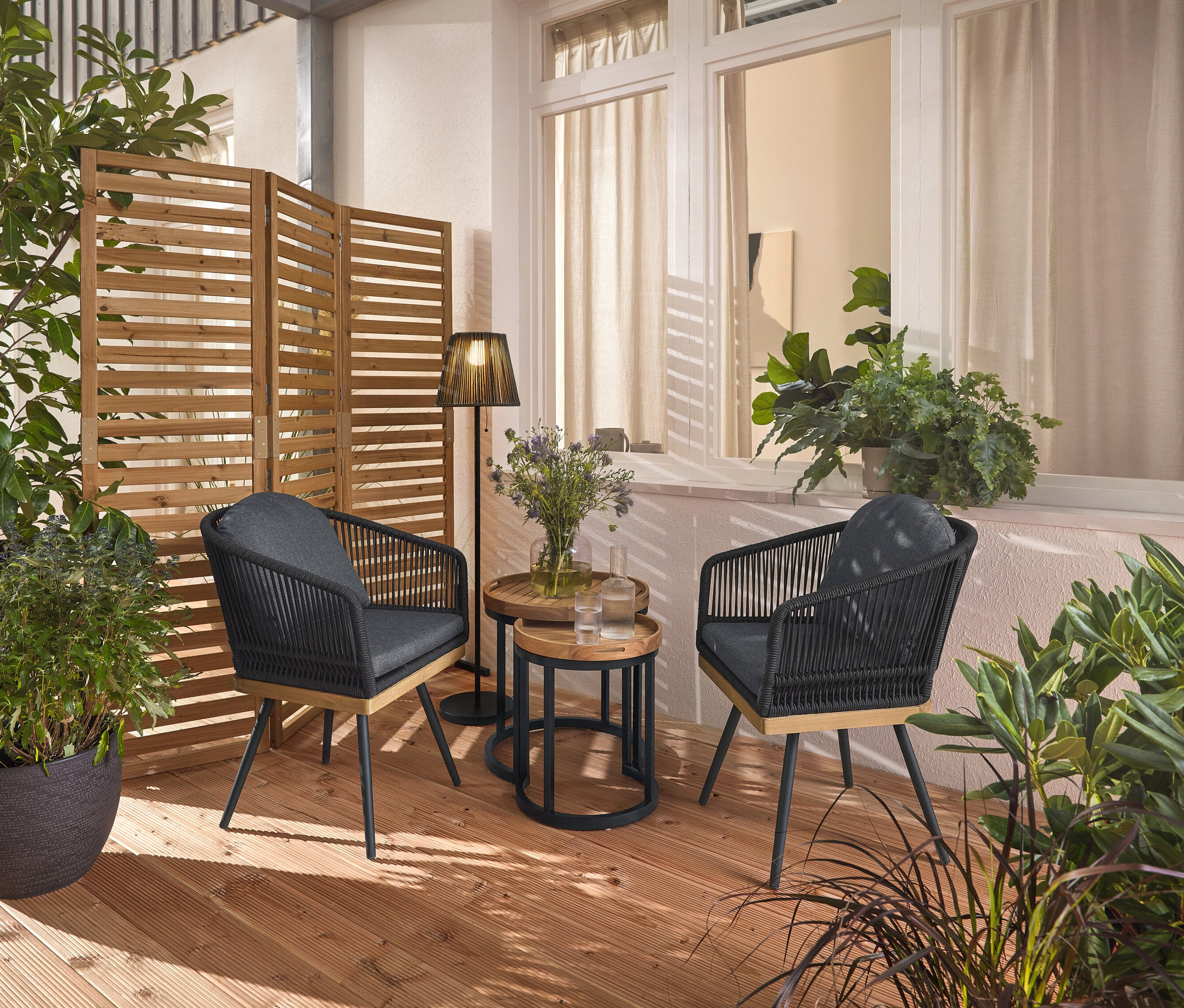 Két forgatható, textilfonatos kerti szék áll egy fa teraszon, köztük két kerti asztal levehető tálcákkal, Ø kb. 40 cm és 50 cm. A háttérben egy rugalmas paraván és egy akkumulátoros napelemes állólámpa.