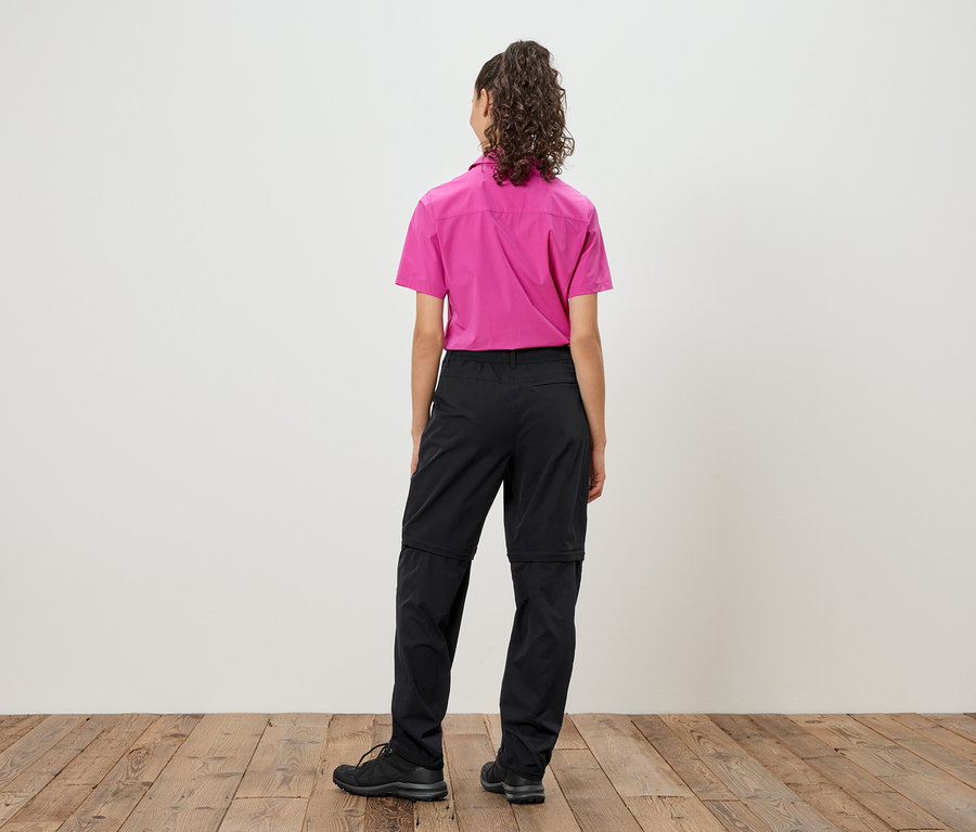 Hátulnézet egy göndör hajú nőről, aki rózsaszín inget és fekete, cipzáras nadrágot visel fekete cipővel.