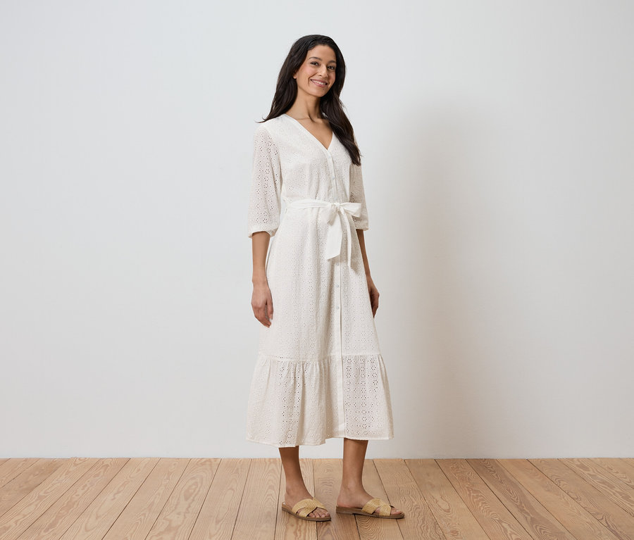 Egy nő áll egy fehér fal előtt, fehér ruhát visel övvel és szandállal.