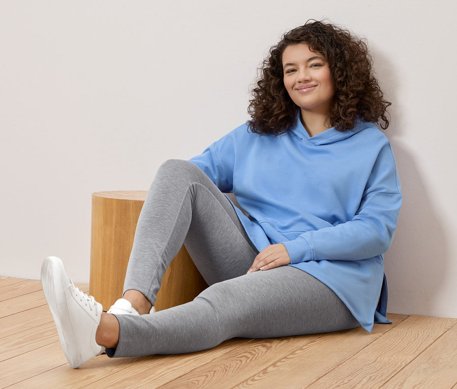 Göndör hajú nő ül a földön, világoskék kapucnis pulóvert, szürke leggingset és fehér tornacipőt visel.