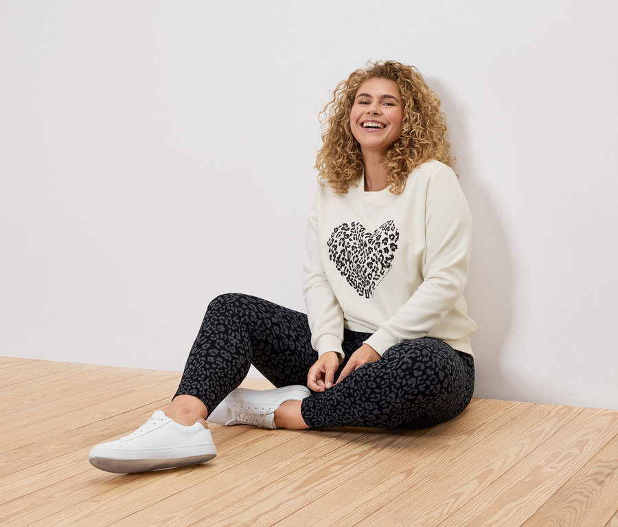 Egy göndör hajú nő ül egy fapadlón, krémszínű pulóvert visel fekete leopárd szívvel, leopárdmintás leggingset és fehér tornacipőt.