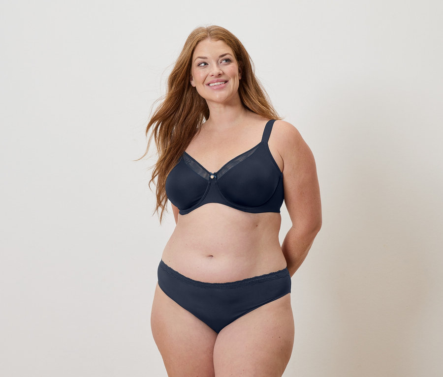 Hosszú vörös hajú nő tengerészkék melltartót és alsóneműt visel, és mosolyog.