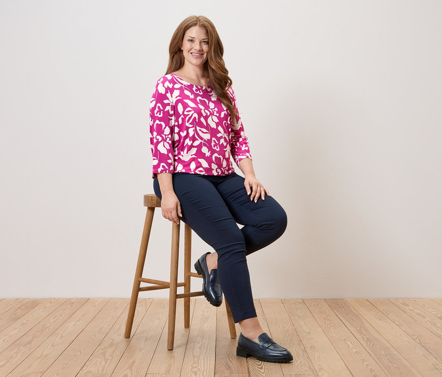 Nő ül egy széken, virágmintás rózsaszín blúzt, kék nadrágot és bőrcipőt visel.