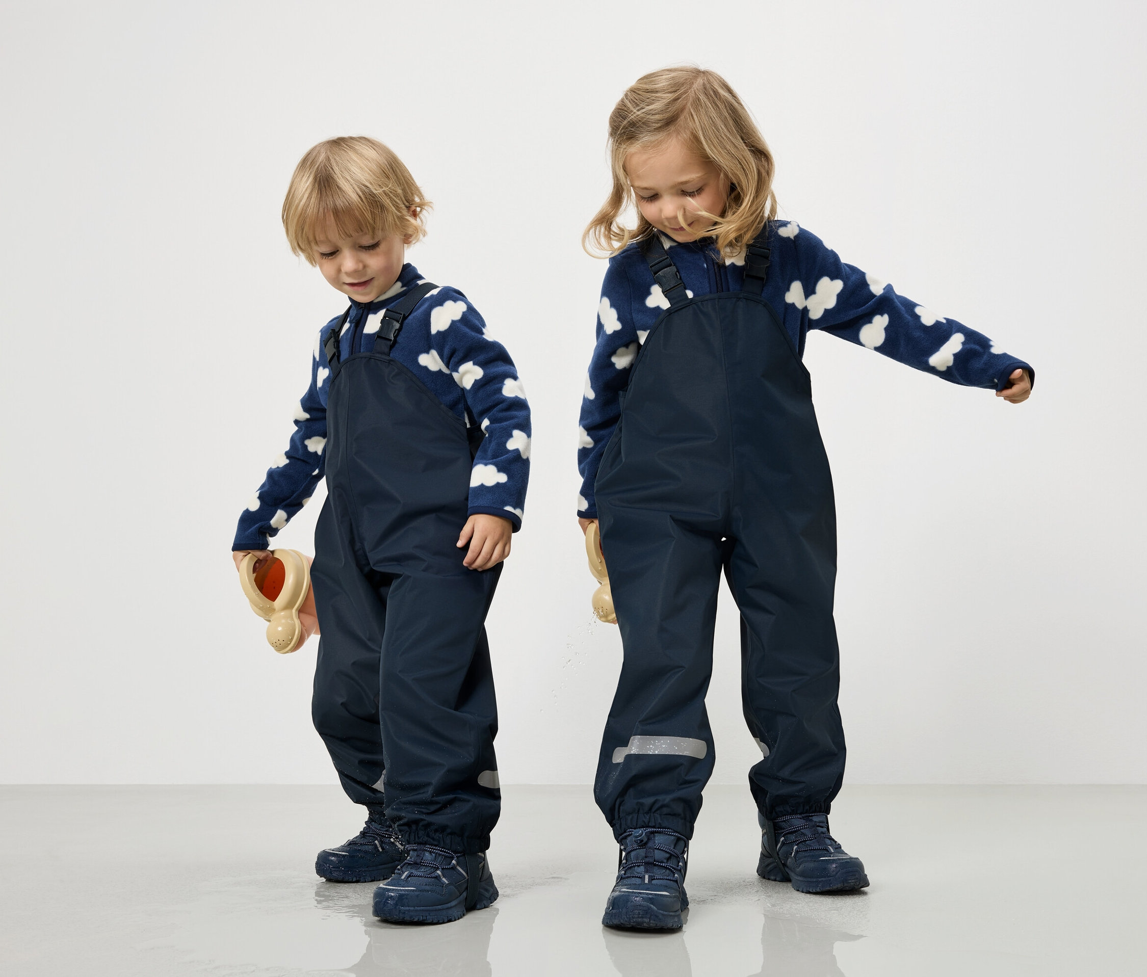 Két gyermek tengerészkékOverallban és kék-fehér felhős pulóverben áll fehér háttér előtt.