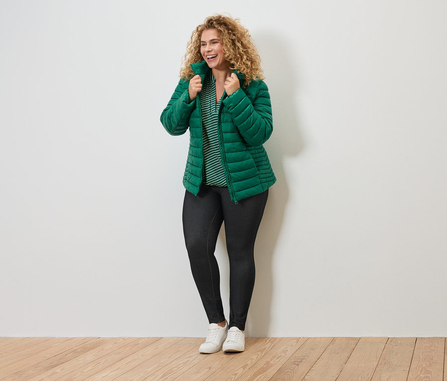 Mosolygó, göndör hajú nő zöld steppelt kabátban, csíkos blúzban, sötét leggingsben és fehér tornacipőben.