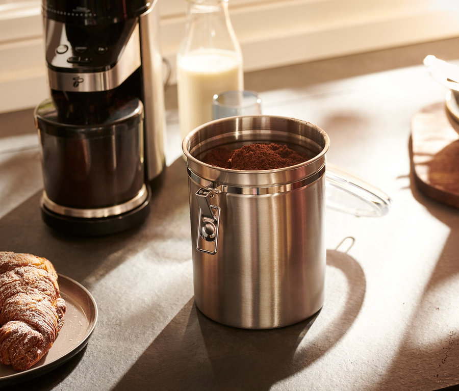 Egy rozsdamentes acélból készült aromatartó doboz áll a konyhapulton egy kávéfőző és egy croissant-t tartalmazó tányér mellett.