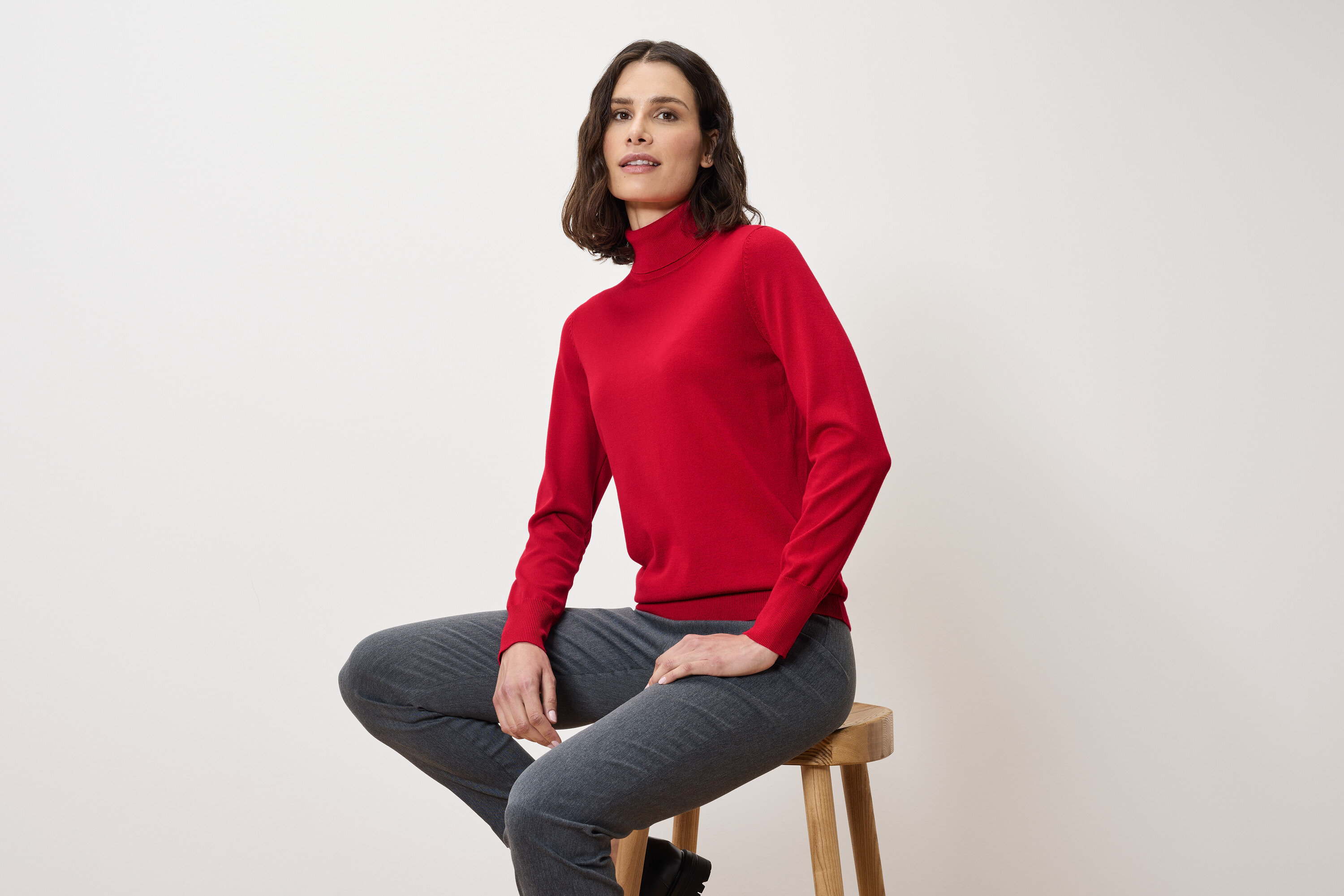 Egy nő ül egy széken, piros garbót és szürke nadrágot visel.