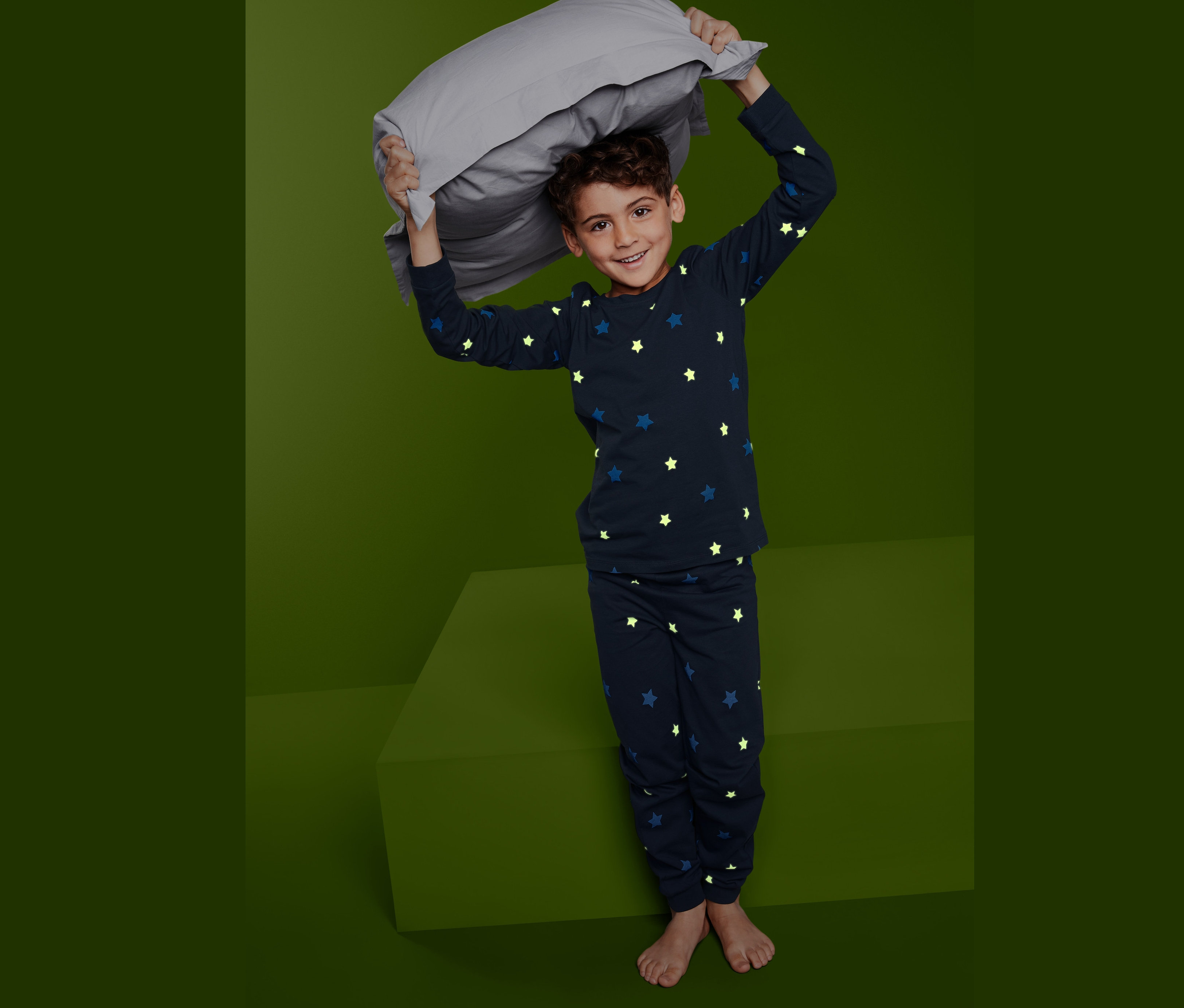 Fiú szürke párnát tart a feje fölött, sötétkék, csillagos Gyerek pizsama, foszforeszkáló  szettben.