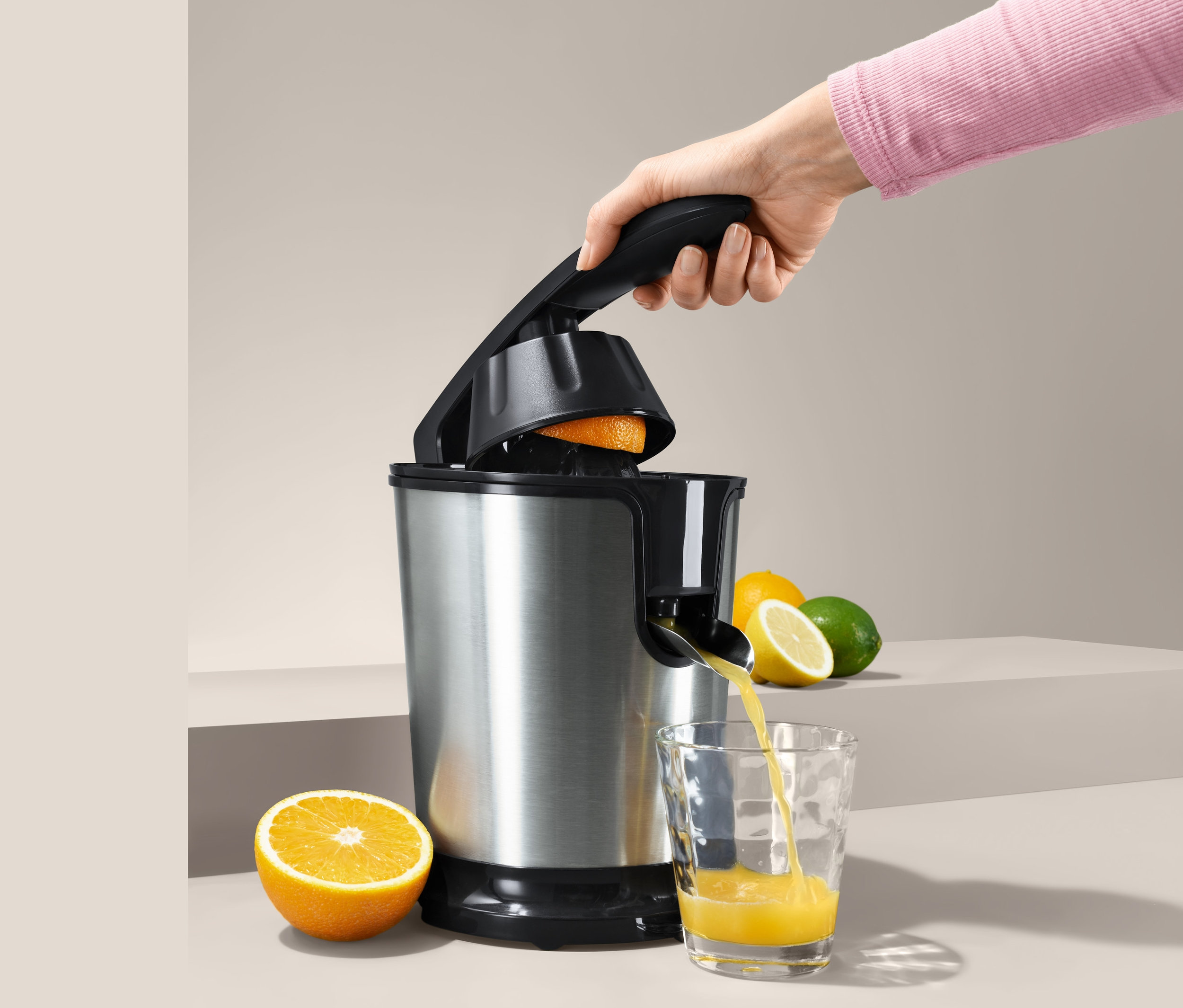 Egy kéz narancsot présel egy elektromos narancsfacsaróban, a lé egy pohárba folyik.