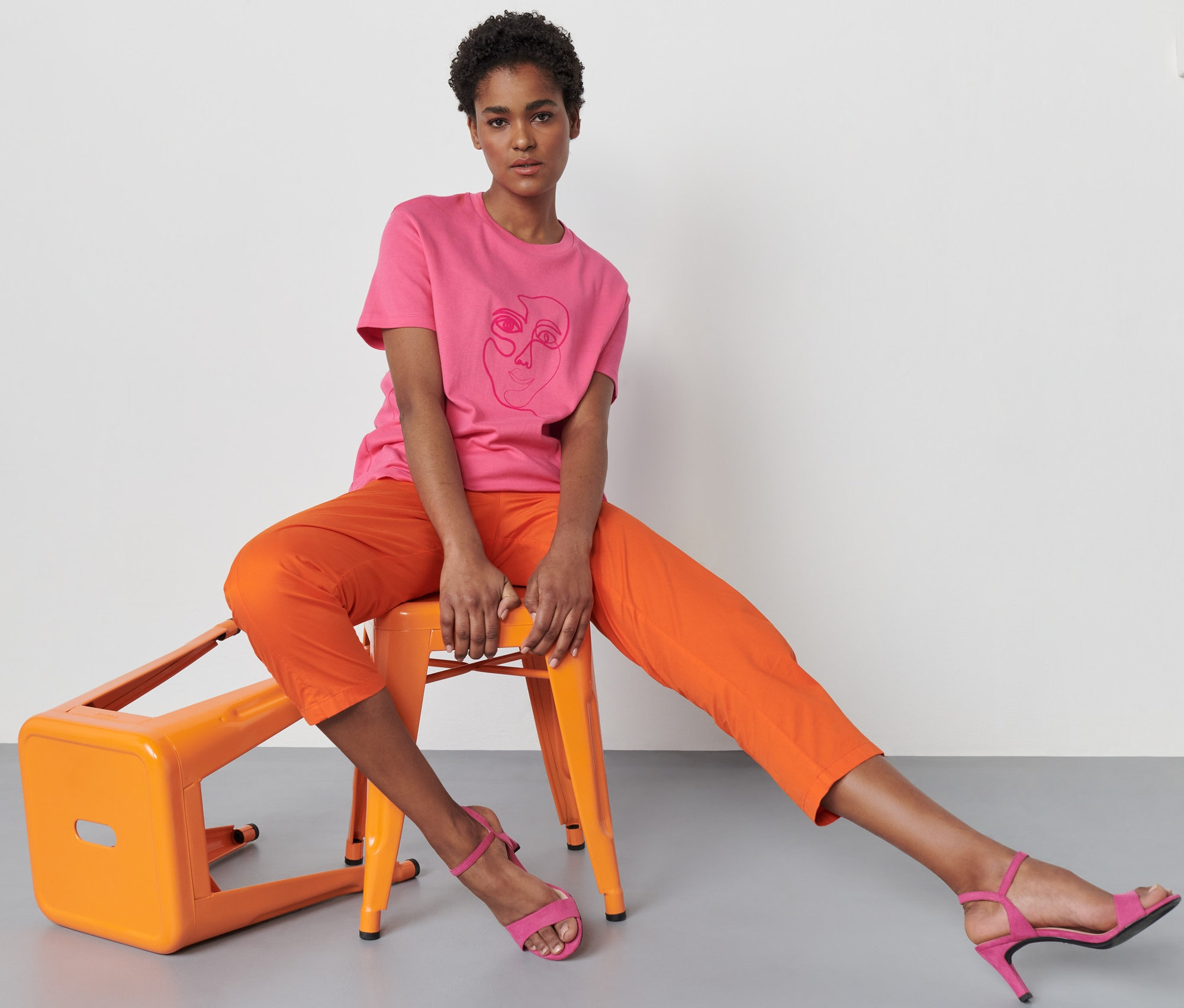 Nő ül egy narancssárga széken, és rózsaszín, nyomott mintás pólót visel.