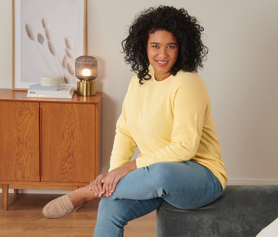 Nő ül egy szürke kanapén, sárga kötött pulóvert, treggingst és velúrbőr mokaszint visel.
