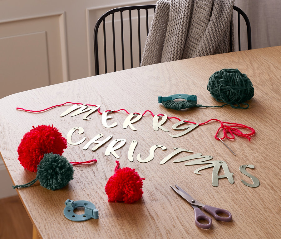 Az asztalon egy MERRY CHRISTMAS girlandkészítő készlet látható. Piros és zöld pomponok, zöld fonal, olló és a "MERRY CHRISTMAS" felirat betűi vannak a képen.
