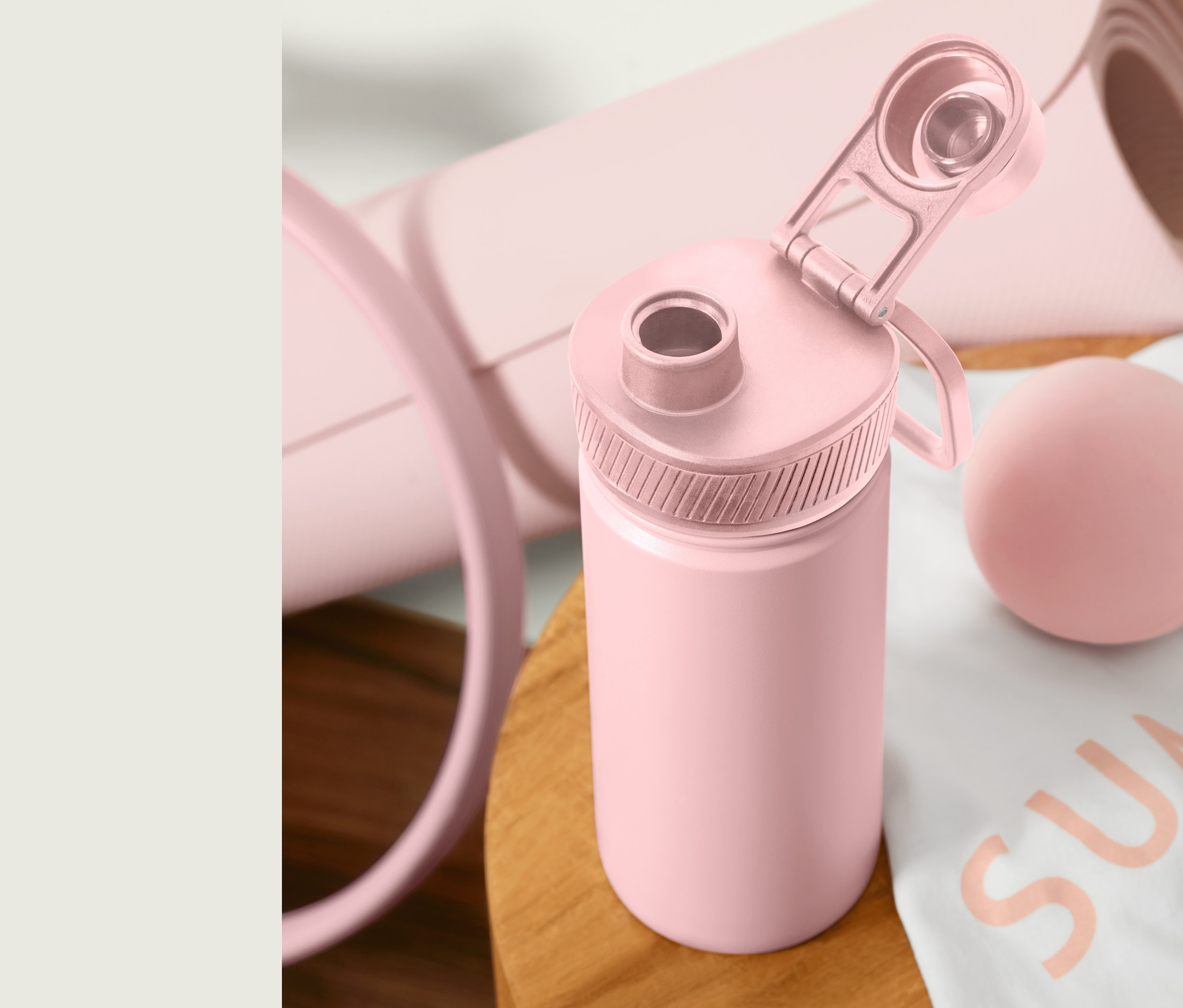 Egy rózsaszín nemesacél kulacs nyitott fedéllel egy faasztalon áll, egy feltekert jógaszőnyeg és egy masszázslabda mellett.