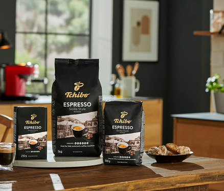 Három csomag Tchibo Espresso Sicilia Style kávé (1 kg, 500 g, 250 g) egy fa asztalon, egy eszpresszó csésze és egy tányér sütemény mellett.