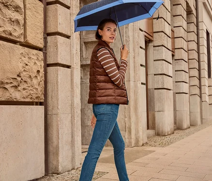 Egy nő sétál az utcán egy esernyővel, Női kifordítható steppelt mellény, sötétkék/barna, Női farmernadrág – Fit »Emma«, sötétkék és Női bőr sneaker cipő, fehér/fekete van rajta.