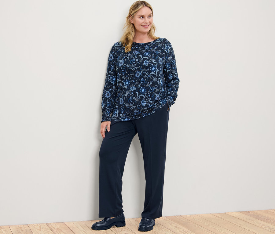 Nő pózol virágos hosszú ujjú pólóblúzban és sötétkék jersey nadrágban.