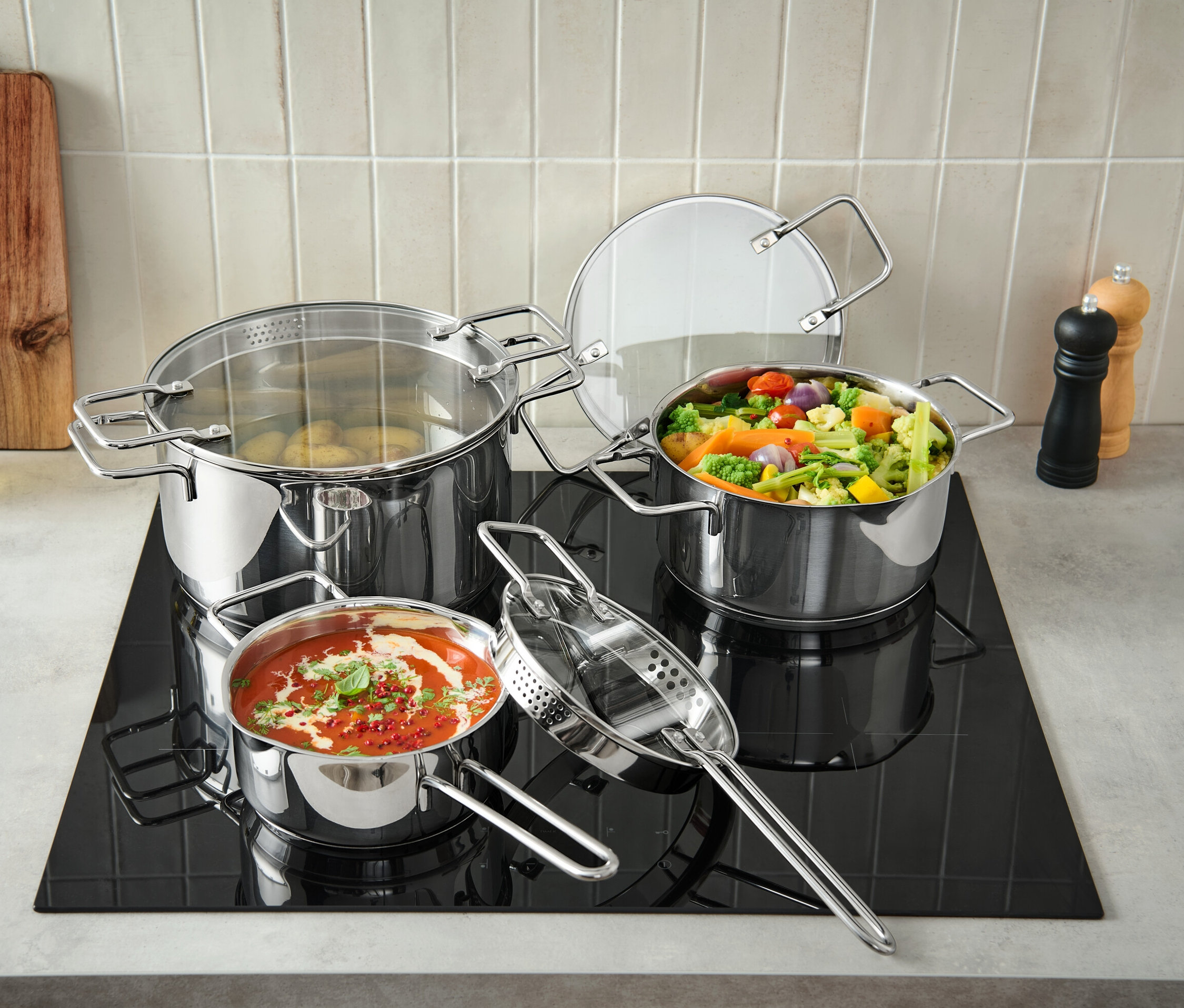 Három darabos, nemesacél edénykészlet a fekete főzőlapon, az egyik edény krumplival, a másik zöldségekkel, a harmadik paradicsomszósszal van tele.