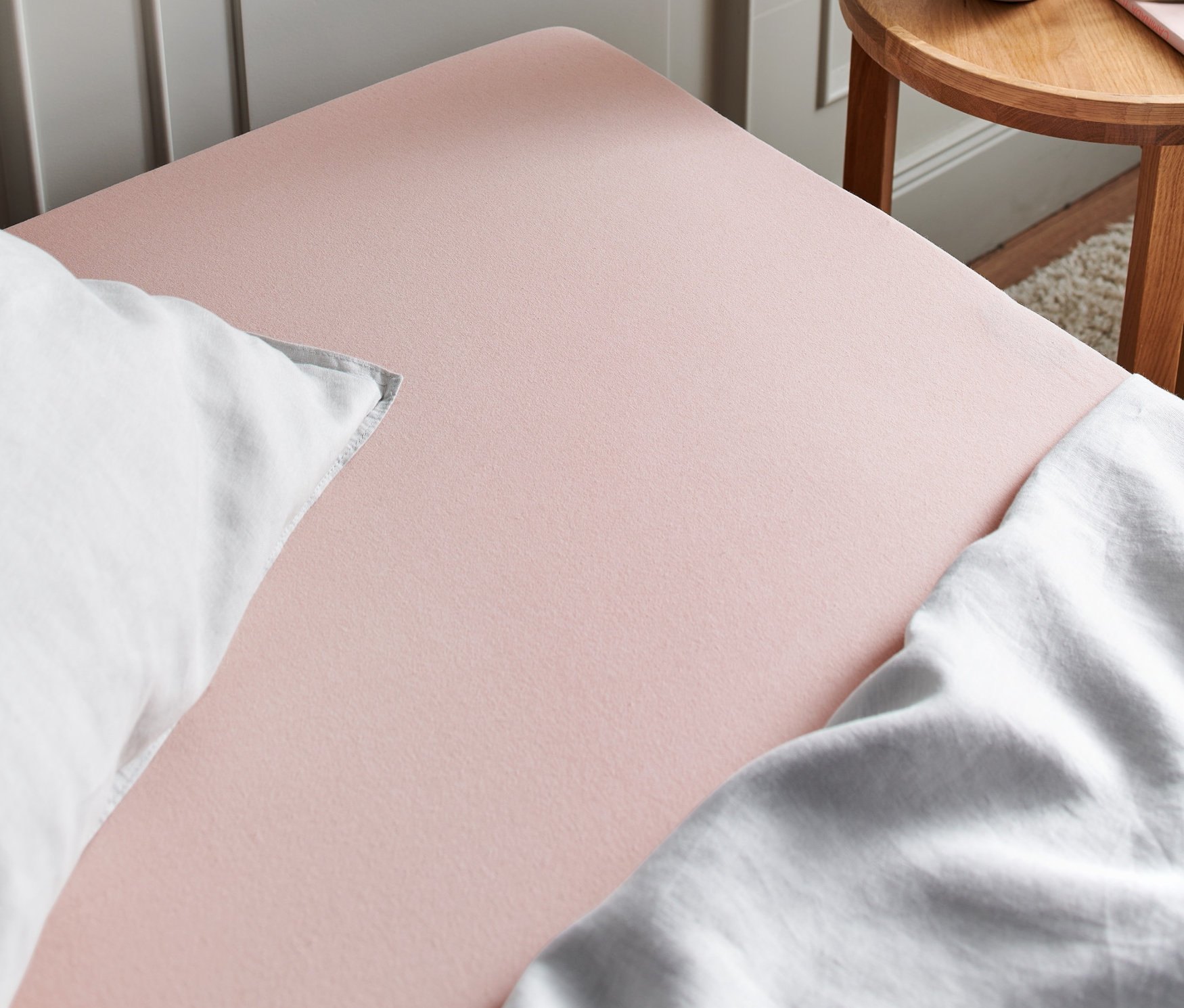 Rózsaszín gumis dzsörzé lepedő normál méretben és egy fehér párna egy ágyon. Az ágy mellett egy asztal található vázával és virágokkal.