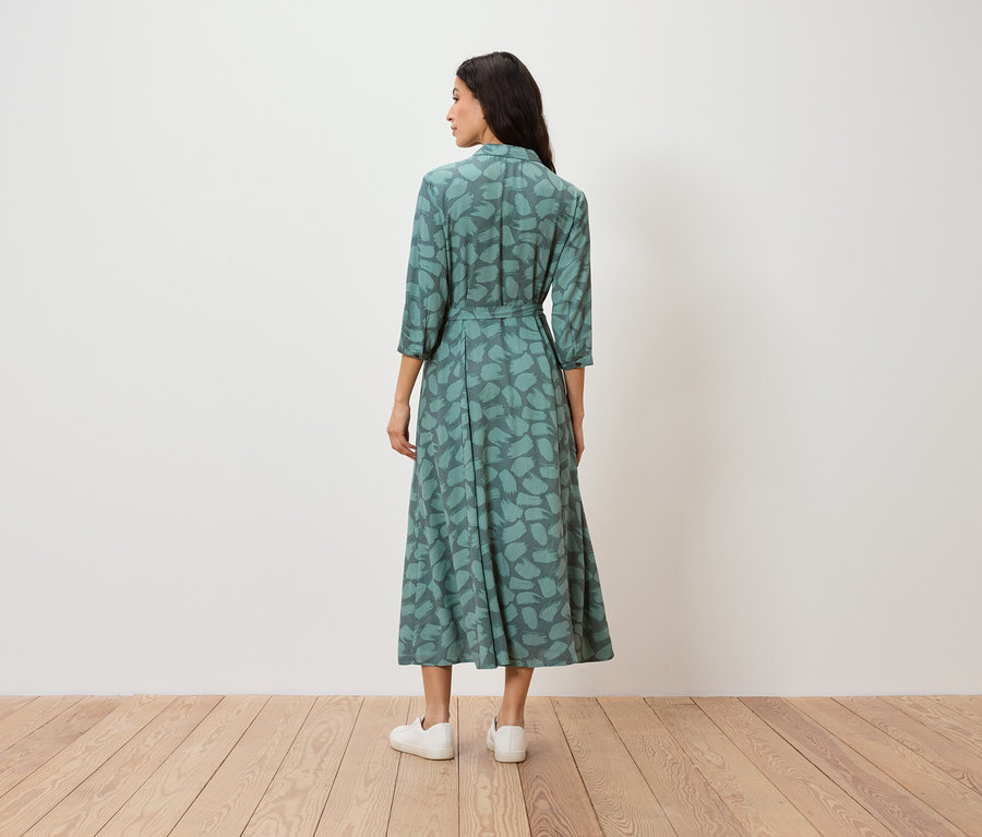 Egy nő áll háttal a kamerának, zöld mintás ruhát és fehér tornacipőt visel.