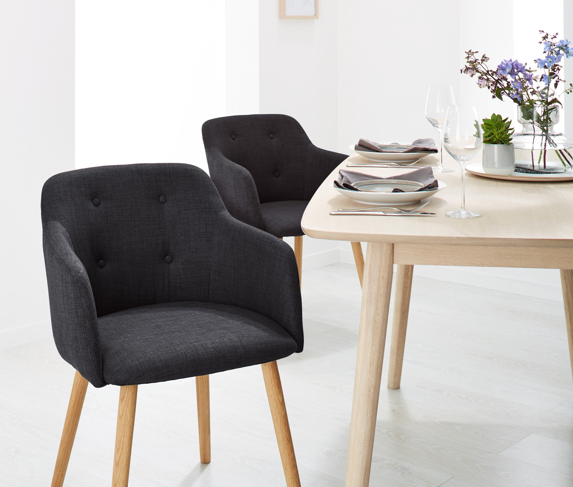 Két antracit színű, kárpitozott szék karfával és egy fa asztal virágokkal és tányérokkal egy étkezőben.