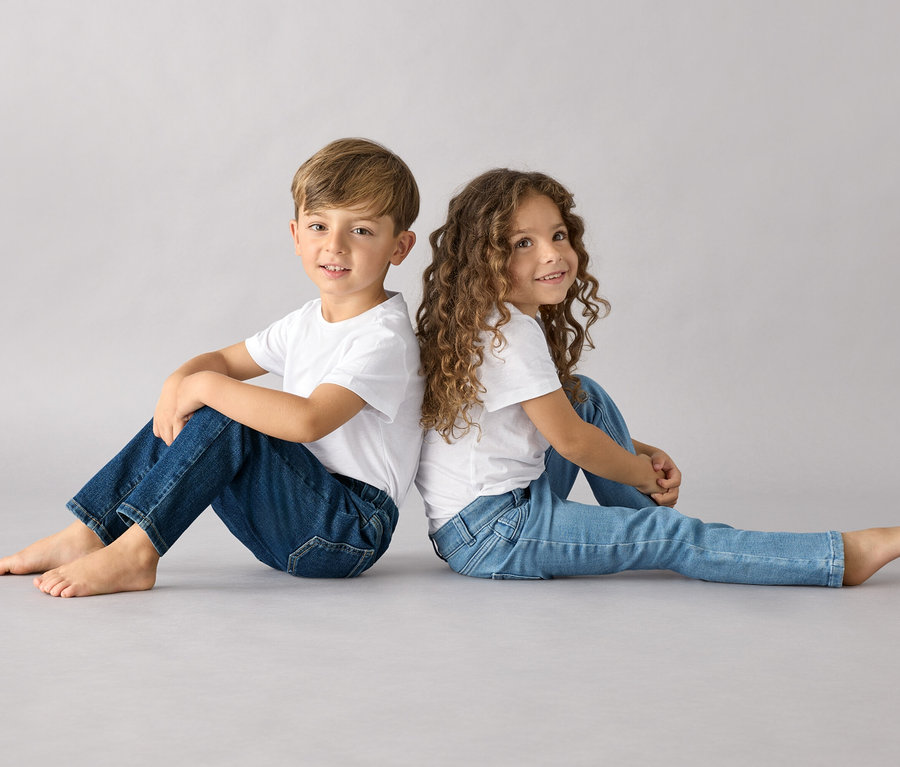 Egy fiú és egy lány háttal ülnek egymásnak, mindketten fehér pólót és Belebújós nadrágot viselnek gyerekeknek – Fit »Luca«.