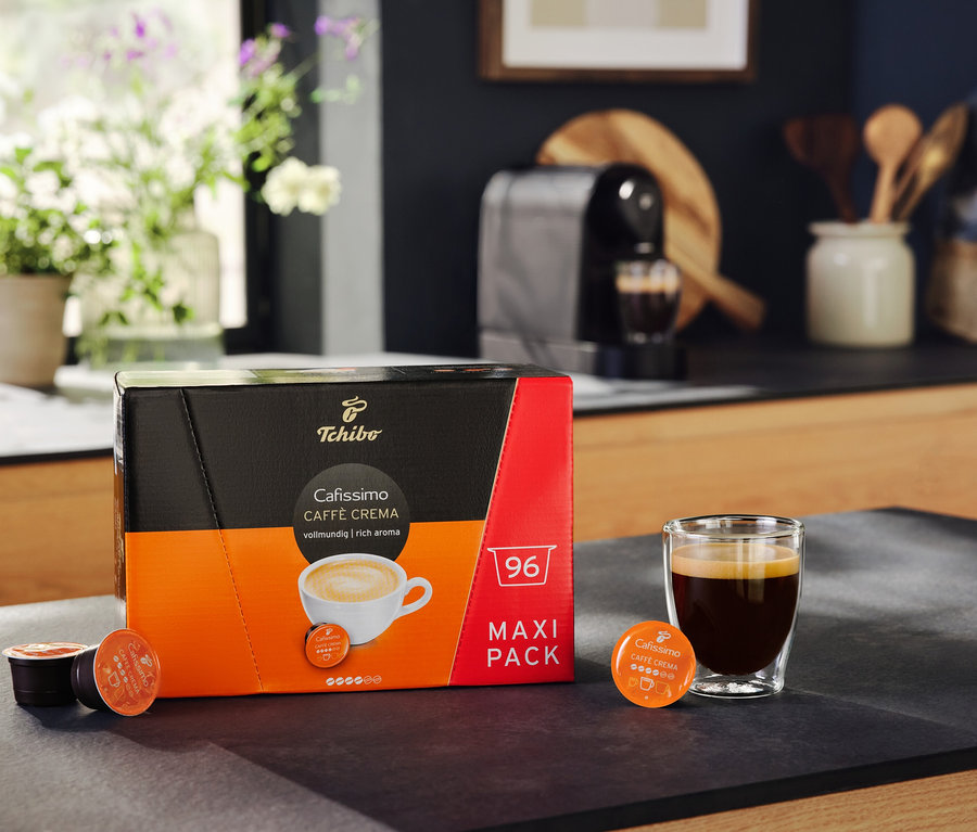 Egy csomag Cafissimo Caffè Crema vollmundig 96 kapszula egy kávés pohár mellett. A háttérben egy Cafissimo pure+ fekete kávéfőző látható.