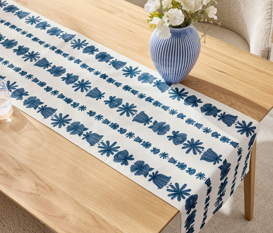 Bézs fa asztal kék-fehér asztali futóval, kék váza fehér virágokkal és egy pohár.