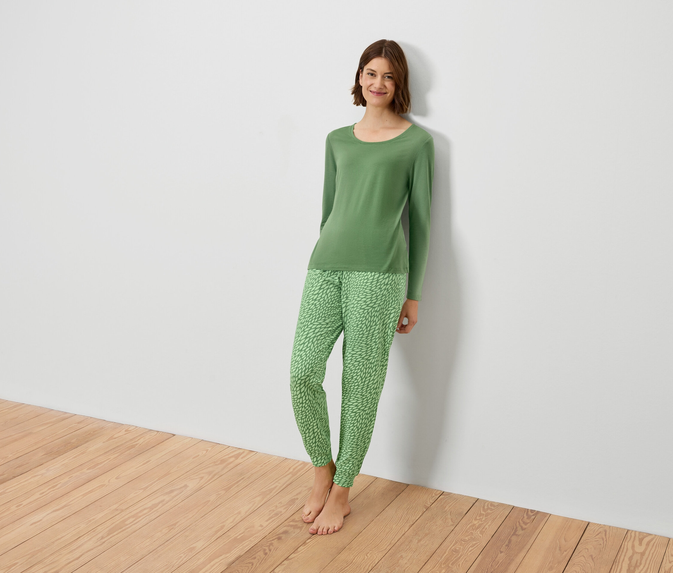 Zöld pizsamában lévő nő támaszkodik egy fehér falnak.