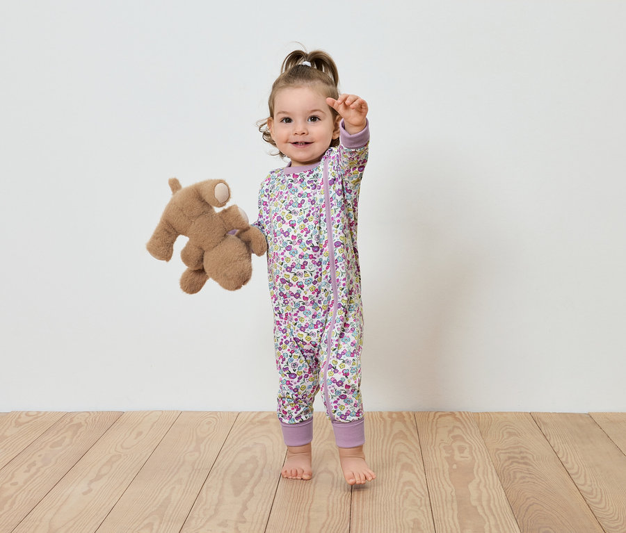 Virágos overallt viselő gyermek áll egy plüssmackóval.