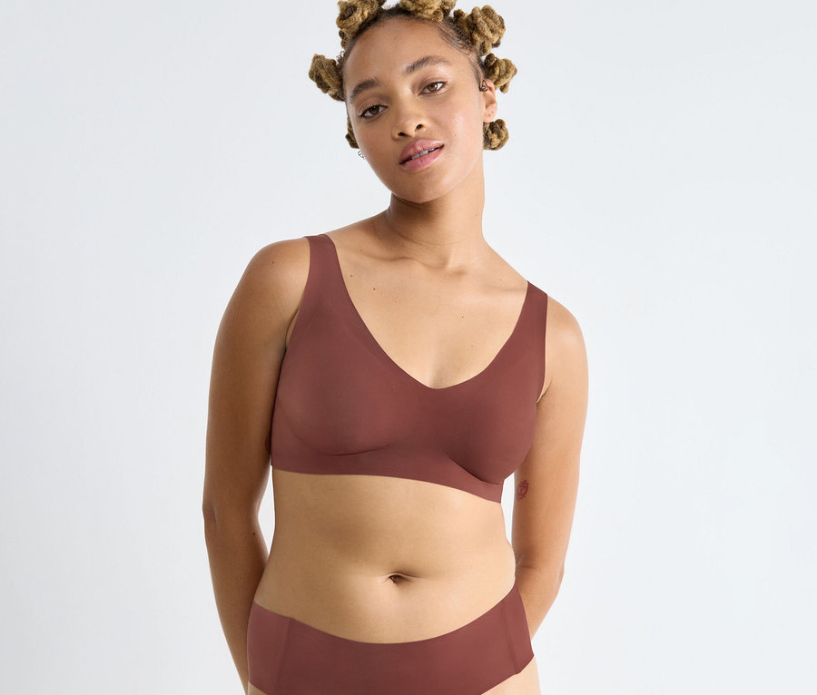 Világosbarna bőrű, raszta hajú nő barna sloggi ZERO Feel 2.0 Bralette melltartót és hozzá illő alsóneműt visel, fehér háttér előtt.
