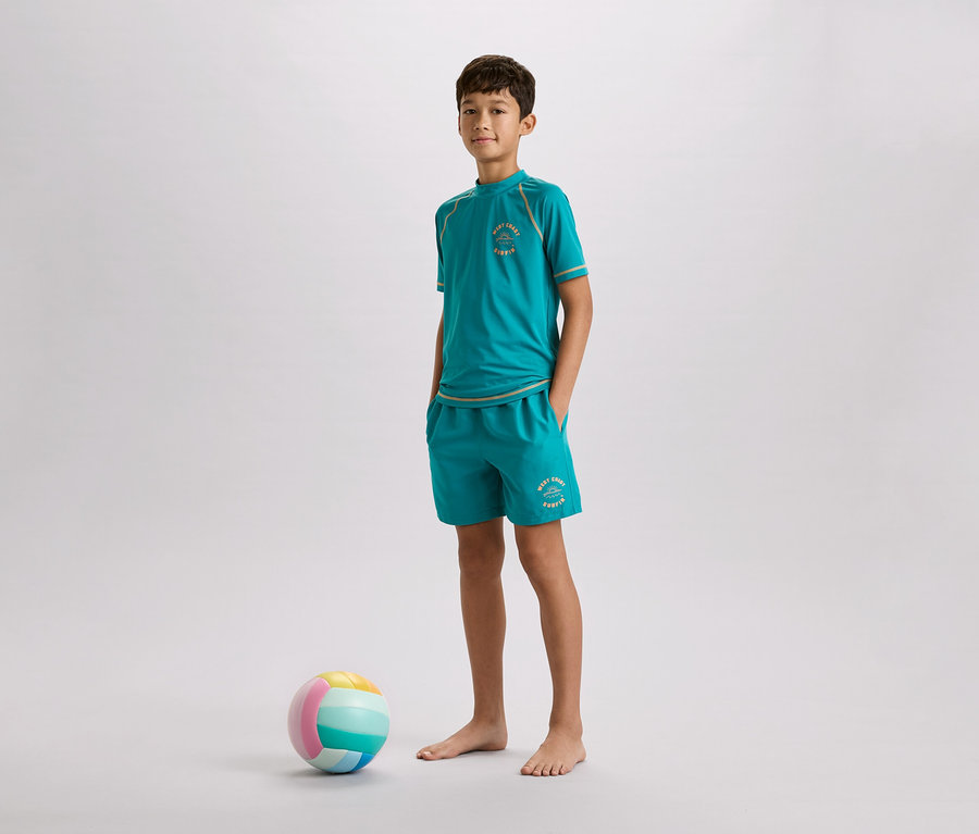 Egy fiú mezítláb áll türkiz pólóban és rövidnadrágban egy színes labda mellett szürke háttér előtt.