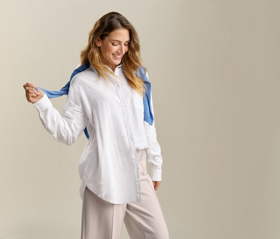 Egy nő kék pulóverrel a vállán hosszú, fehér vászonblúzt visel.