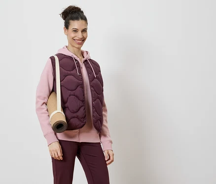 Nő visel bordó steppelt mellényt rózsaszín kapucnis pulóver és bordó leggings felett, a vállán egy jógamatraccal.