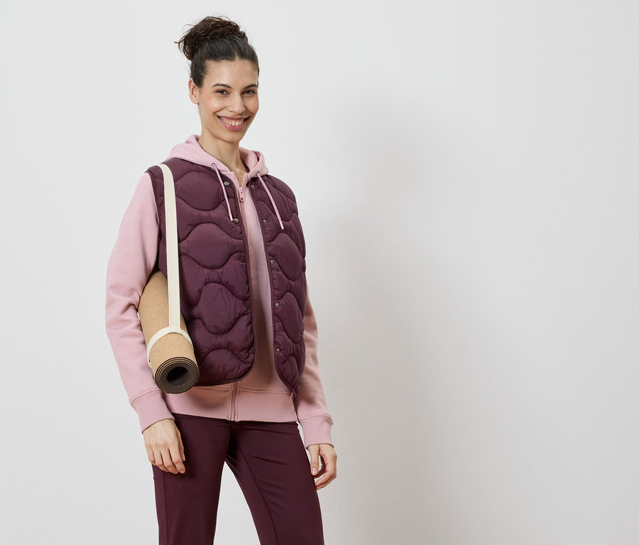 Nő visel bordó steppelt mellényt rózsaszín kapucnis pulóver és bordó leggings felett, a vállán egy jógamatraccal.