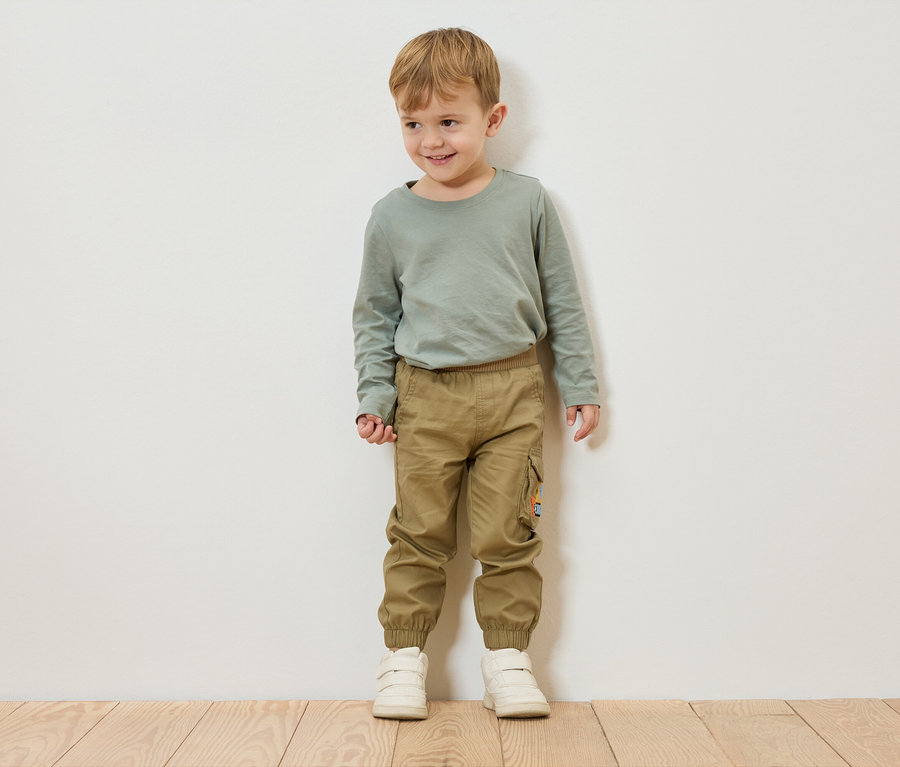 Egy szőke hajú fiú áll egy fehér fal előtt, zöld hosszú ujjú pólót, khaki nadrágot és fehér cipőt visel.