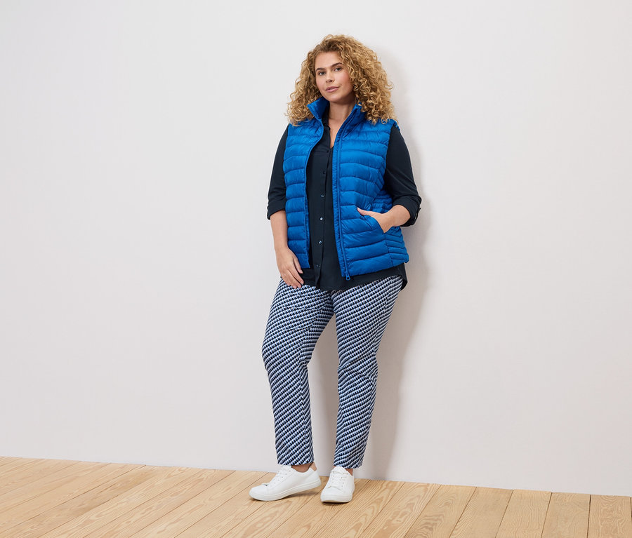 Göndör hajú nő kék mellényben, sötét ingben és mintás nadrágban áll egy fehér falnál.