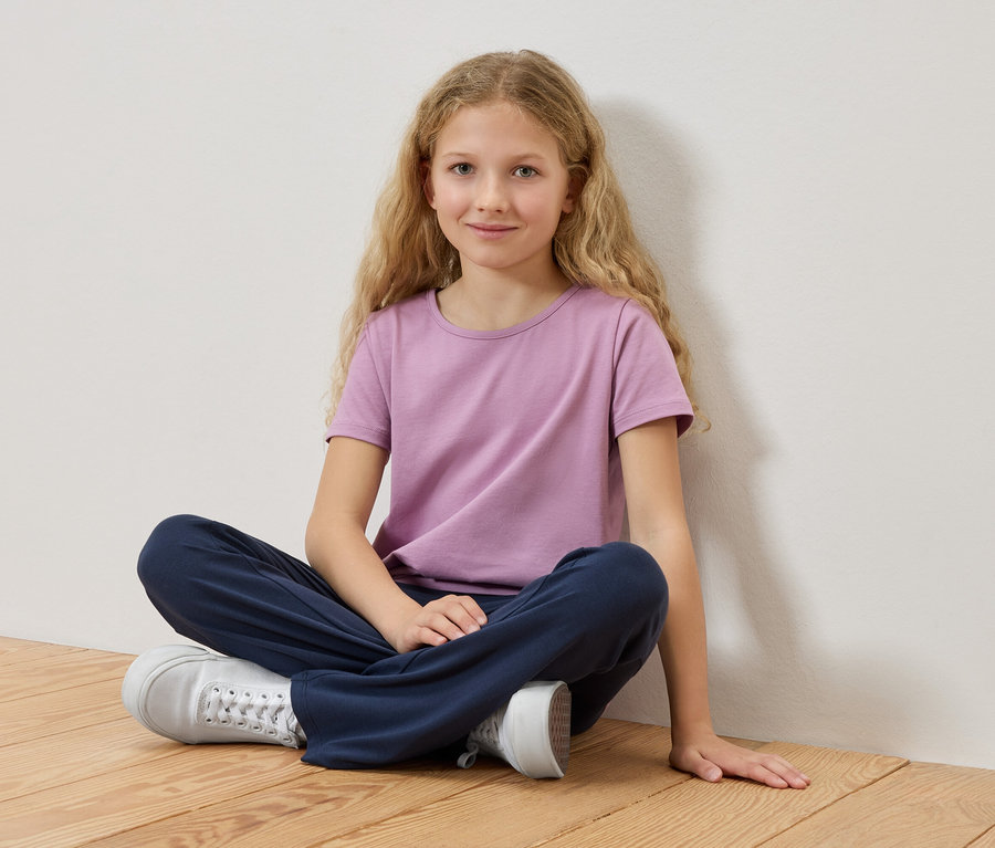 Egy szőke hajú lány ül keresztbe tett lábakkal egy fapadlón, lila pólót és kék nadrágot visel.