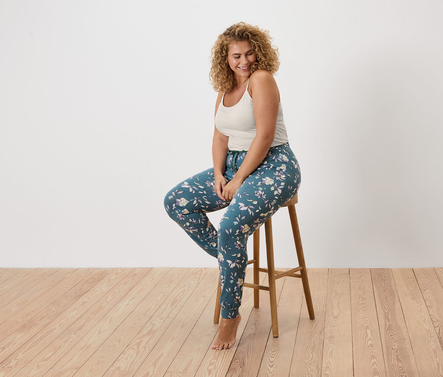 Göndör hajú nő ül egy széken, trikót és virágos leggingst visel.