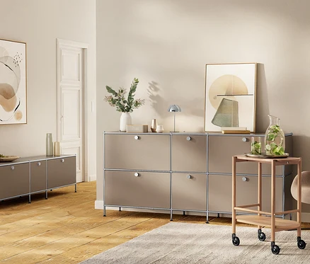 Modern nappali taupe színű bútorokkal. Balra egy »CN3« alacsony komód, 3 lenyíló ajtóval, rajta vázák. Jobbra egy »CN3« nagy fém szekrény, 6 lenyíló ajtóval, rajta »CN3« LED-es asztali lámpa, zöld növényes váza és kép. Mellette egy fa tálalókocsi kancsóval és poharakkal.