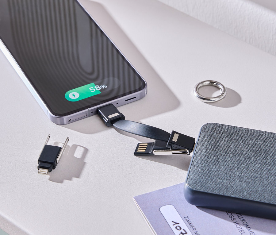 Egy okostelefon töltődik egy kábellel és egy szürke akkumulátorral egy fehér asztalon. Egy gyűrű és egy szobakártya van mellette.