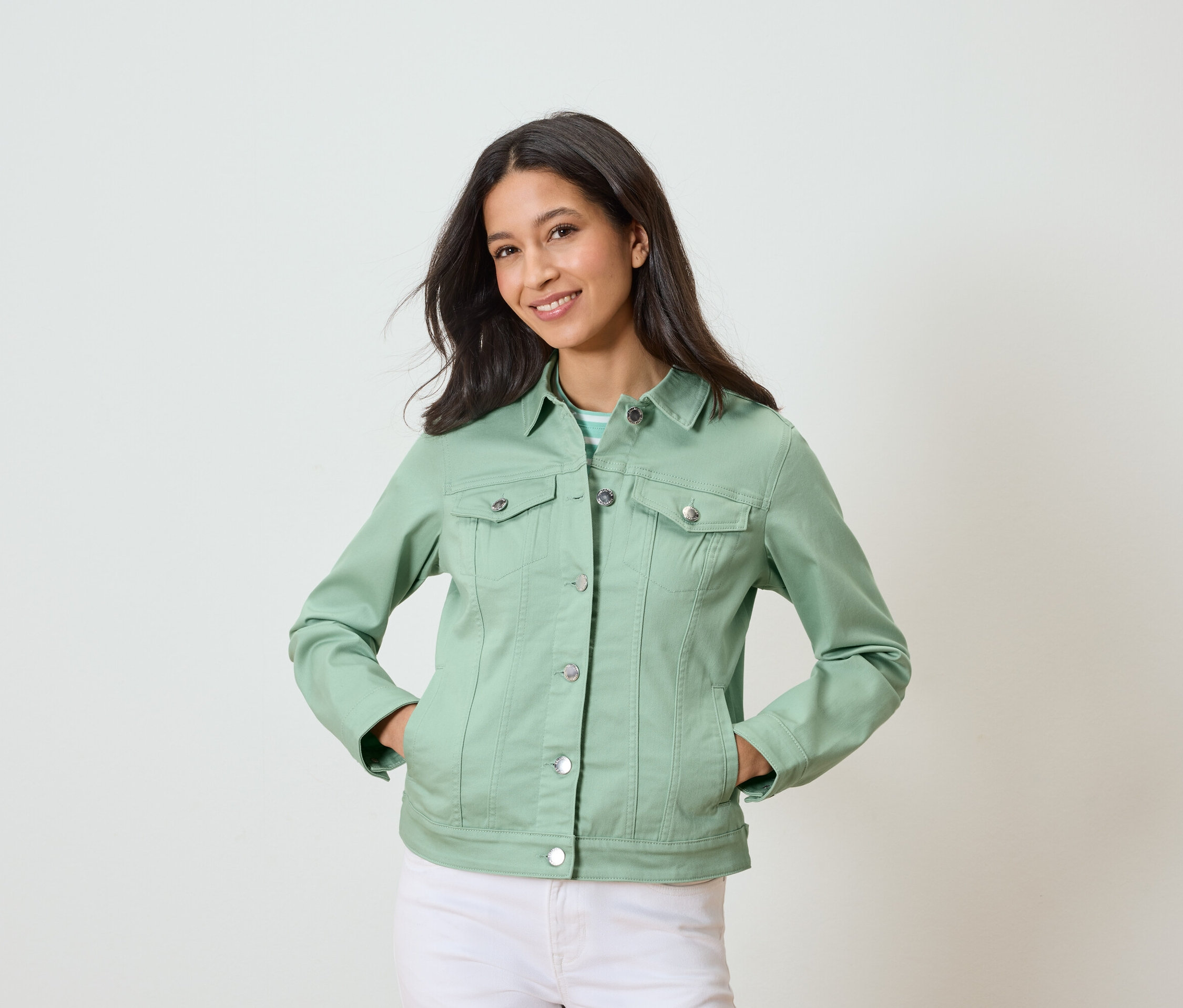 Nő pózol zsebre tett kézzel, világoszöld farmerdzsekiben és fehér nadrágban.