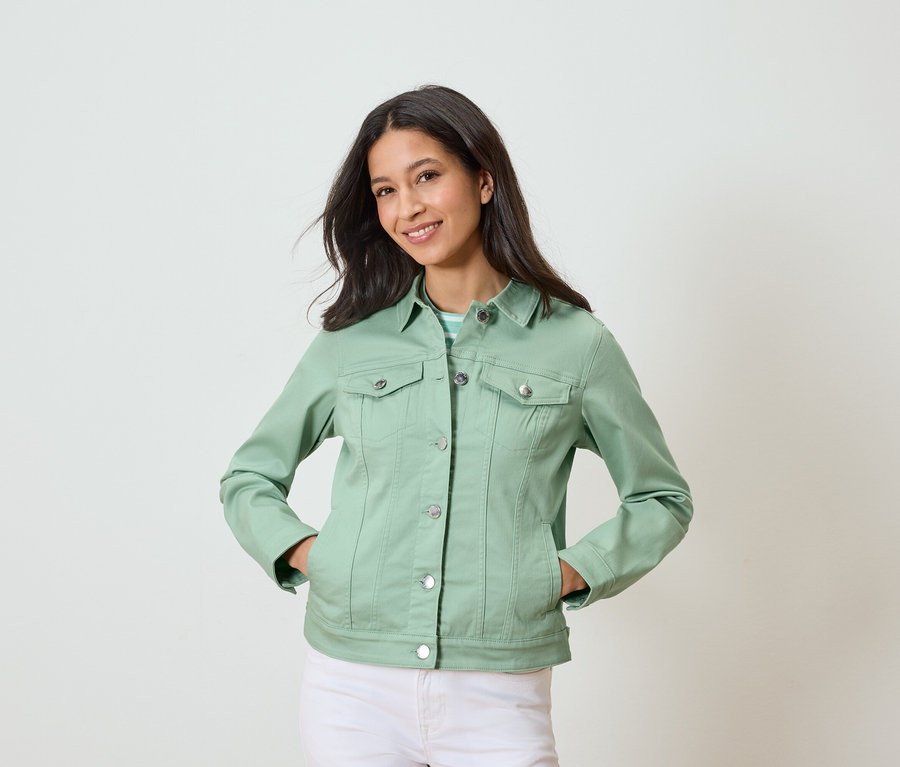 Nő pózol zsebre tett kézzel, világoszöld farmerdzsekiben és fehér nadrágban.