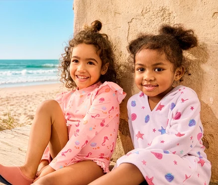 Két lány ül a strandon, az egyiken Kinder-Badeanzug mit UV-Schutzfaktor 50+ és Kinder-Badeschuhe, a másikon Leichter Kinder-Frottierponcho van.