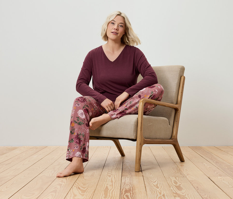 Egy szőke nő ül egy fotelben, bordó hosszú ujjú pólót és virágmintás pizsama nadrágot visel.