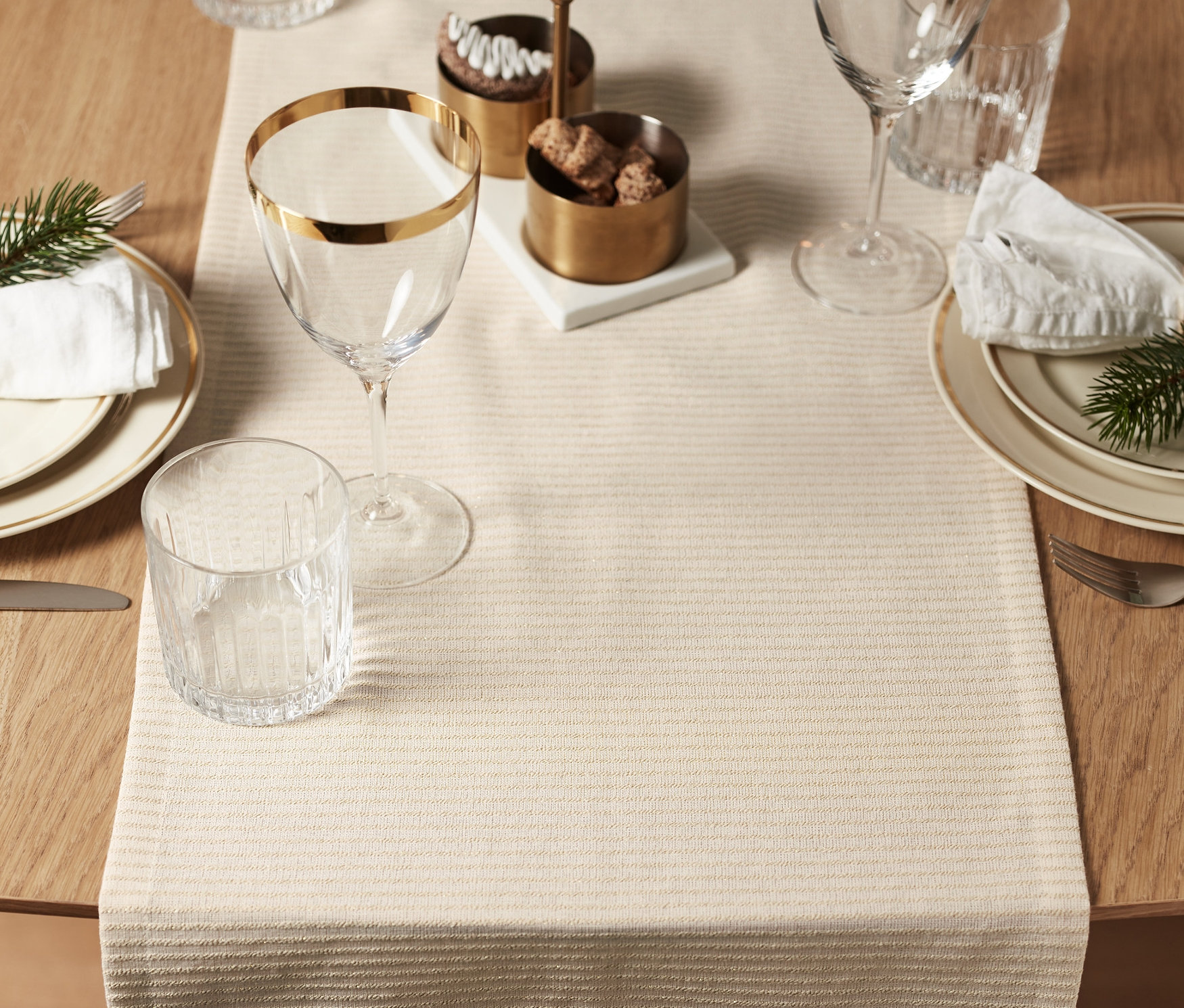 Megterített asztal bézs színű csíkos asztali futóval, tányérokkal, arany szegélyű poharakkal és dekorációkkal.